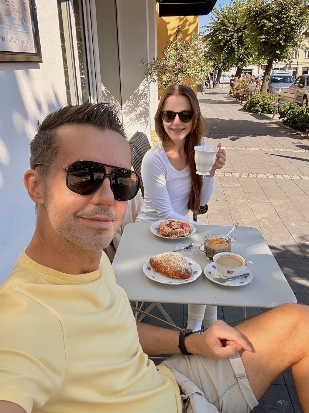 Dwoje ludzi przy stoliku na chodniku przed piekarnią, na blacie dwa wypieki na talerzykach, filiżanka americano i caffè latte w szklance, w tle drzewa i pierzeja rynku
