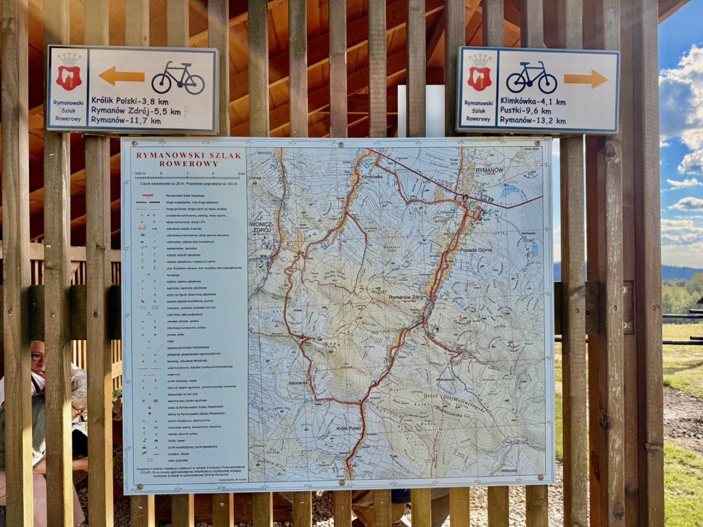 Dwie niebieskie tablice kierunkowe Rymanowskiego Szlaku Rowerowego i plansza z mapą, przykręcone do ściany wiaty z desek
