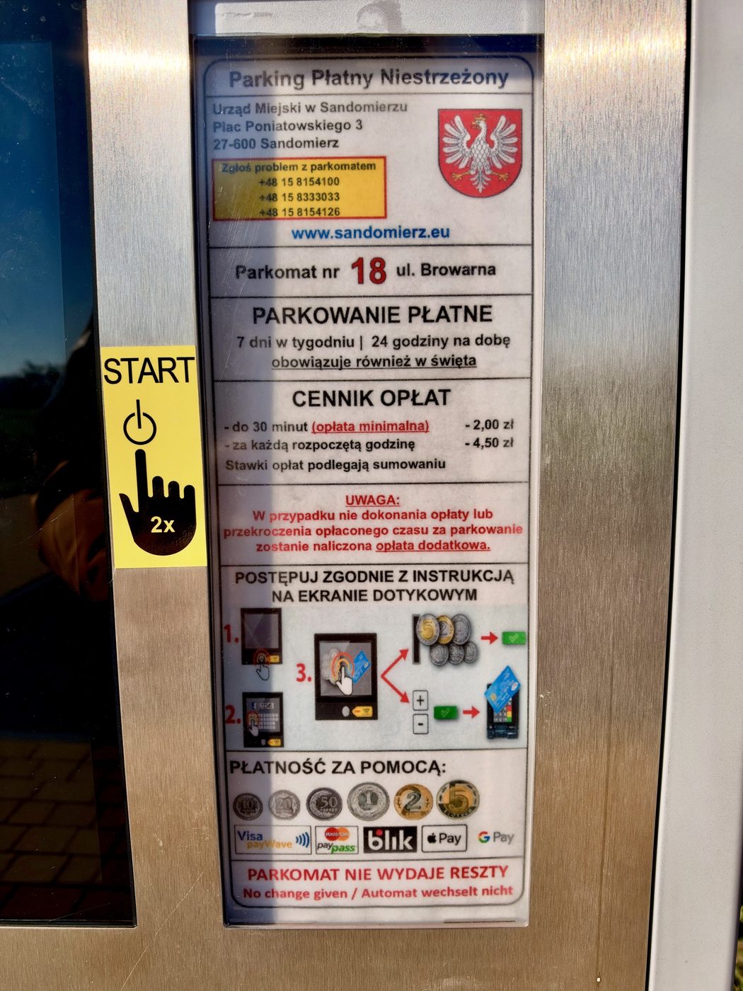 Tabliczka na srebrnym parkomacie: „Parking Płatny Niestrzeżony”, Urząd Miejski w Sandomierzu z herbem, „Parkomat nr 18 ul. Browarna”, „Parkowanie płatne 7 dni w tygodniu, 24 godziny na dobę, obowiązuje również w święta”, cennik opłat, instrukcja obsługi ekranu dotykowego, znaki płatności monetami, kartą, BLIK, Apple Pay i Google Pay oraz napis „Parkomat nie wydaje reszty”, obok żółta naklejka „START 2x”
