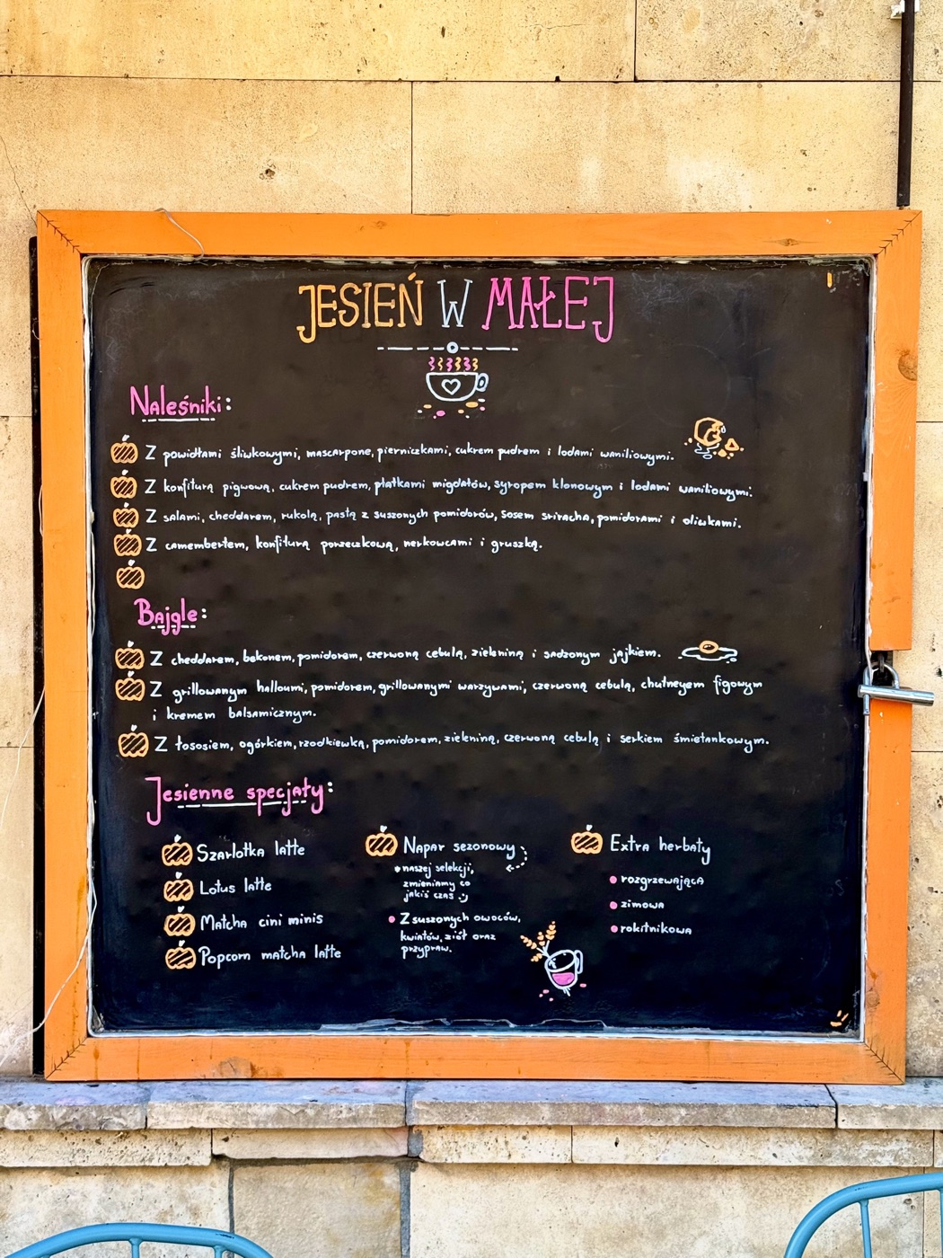 Czarna tablica kredowa w pomarańczowej ramie z ręcznie wypisanym jesiennym menu naleśników, bajgli i kaw, z rysunkami dyni i filiżanki, na ścianie z jasnego kamienia
