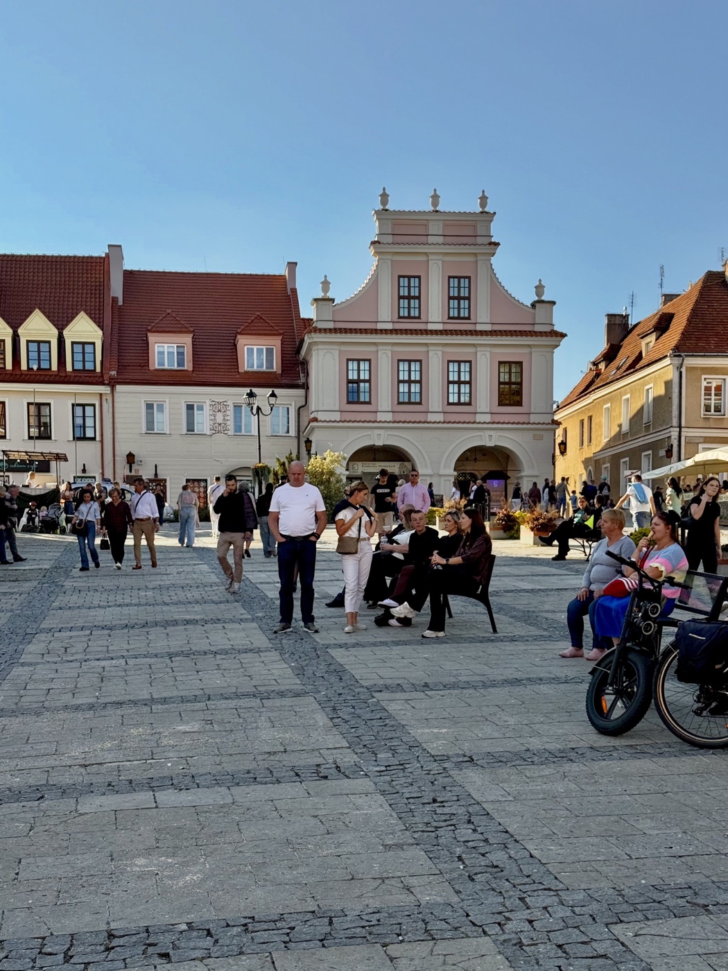Różowo-biała kamienica z dwoma łukami podcieni i barokowym szczytem zwieńczonym kamiennymi wazonami, obok kamienice z czerwonymi dachami i lukarnami, na płycie rynku spacerowicze i ludzie na ławkach
