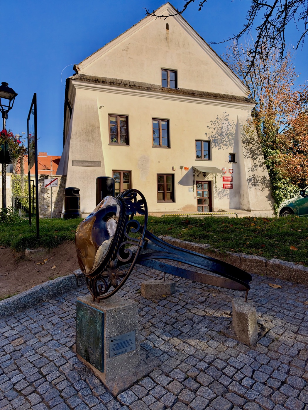 Duży stalowy pierścień leżący na boku, z kutą oprawą i osadzoną w niej błyszczącą brązową bryłą krzemienia, oparty na kamiennym cokole z dwiema tablicami i na dwóch kamiennych bloczkach, na placu z kostki, w tle jasny piętrowy budynek z wysokim szczytem, skarpami przy narożnikach i czerwonymi tablicami przy drzwiach, obok latarnia i drzewo w jesiennych liściach