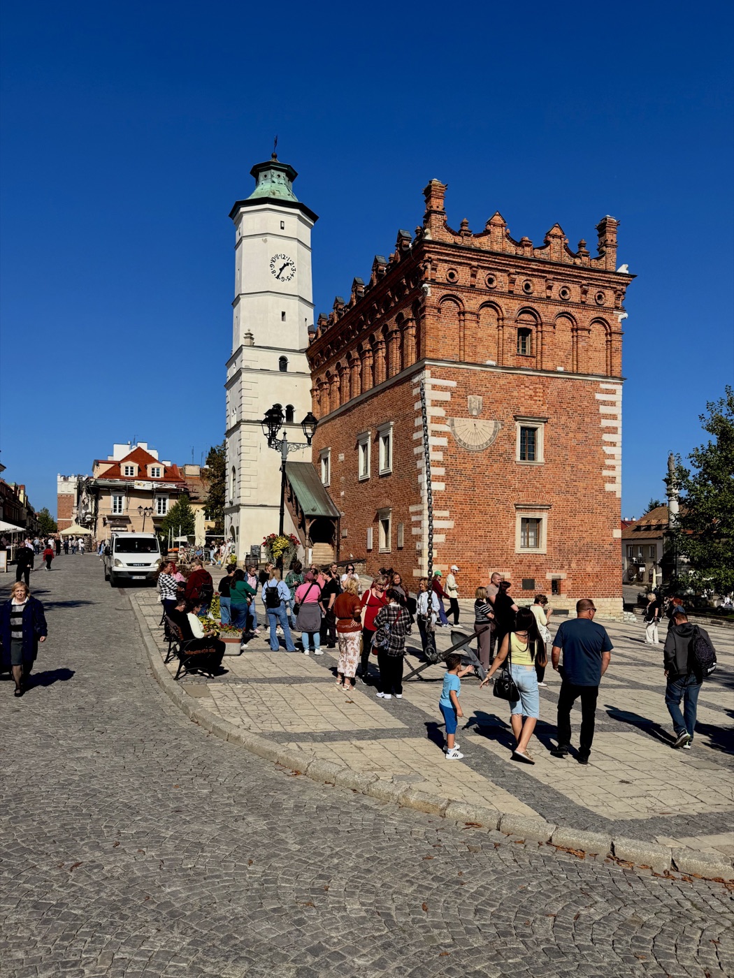 Ceglany ratusz z ząbkowaną attyką i białą wieżą z zegarem, na ścianie zegar słoneczny z herbem, przed narożnikiem kotwica z łańcuchem wzniesionym pionowo w górę, przed ratuszem grupa turystów i biały samochód dostawczy