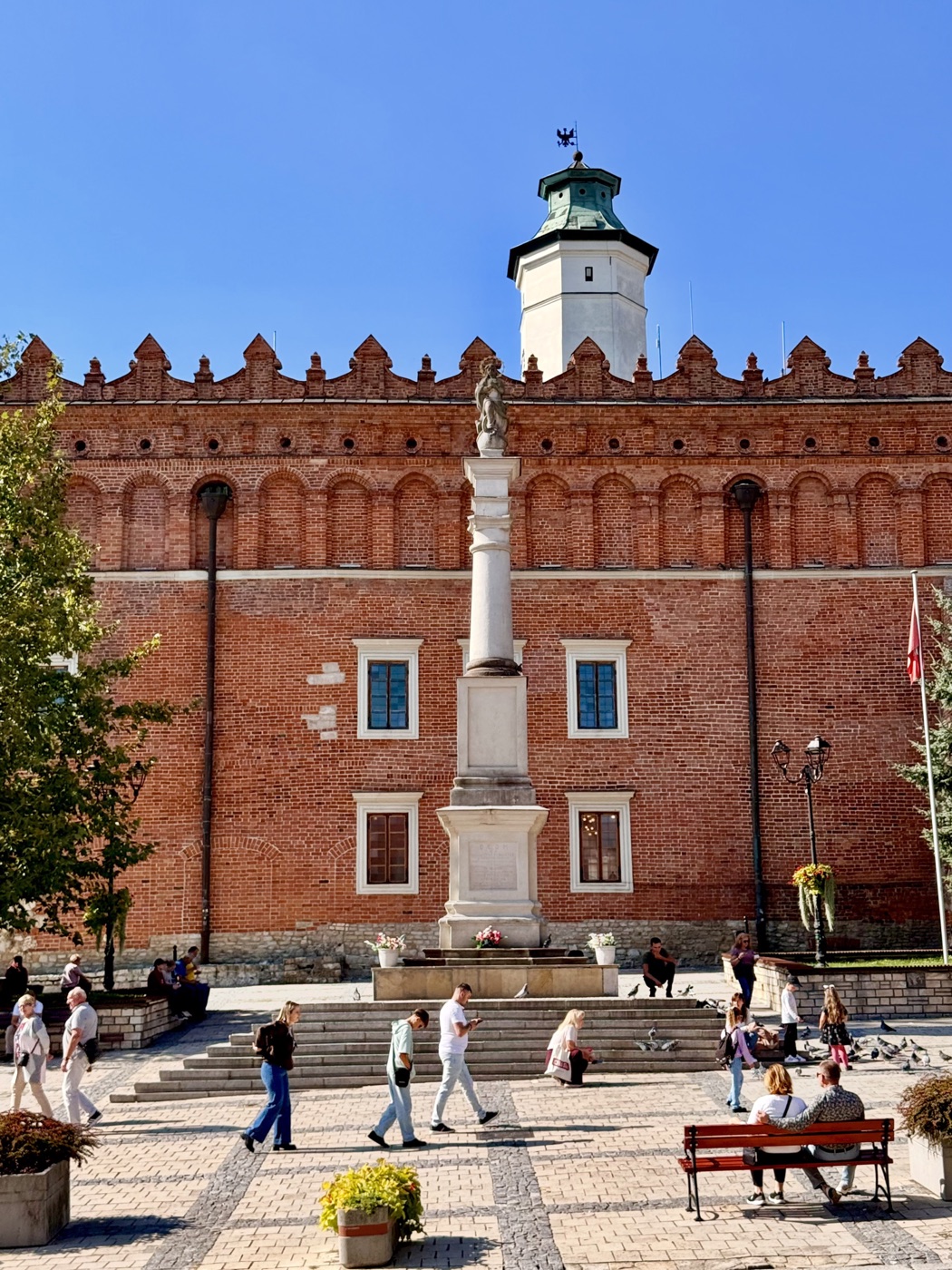 Ceglany ratusz z renesansową attyką i białą ośmioboczną wieżą z zielonym hełmem, przed nim kamienna kolumna z kobiecą figurą, schody, donice z kwiatami i ludzie wśród gołębi