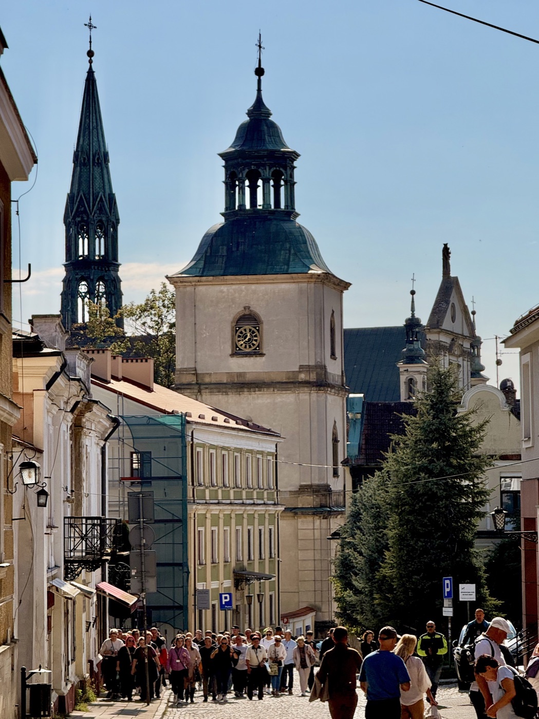 Brukowana ulica Starego Miasta z wolnostojącą wieżą z zegarem i hełmem z latarnią, z lewej zielona iglica, z prawej barokowy szczyt świątyni, kamienice i grupa turystów idąca w stronę aparatu