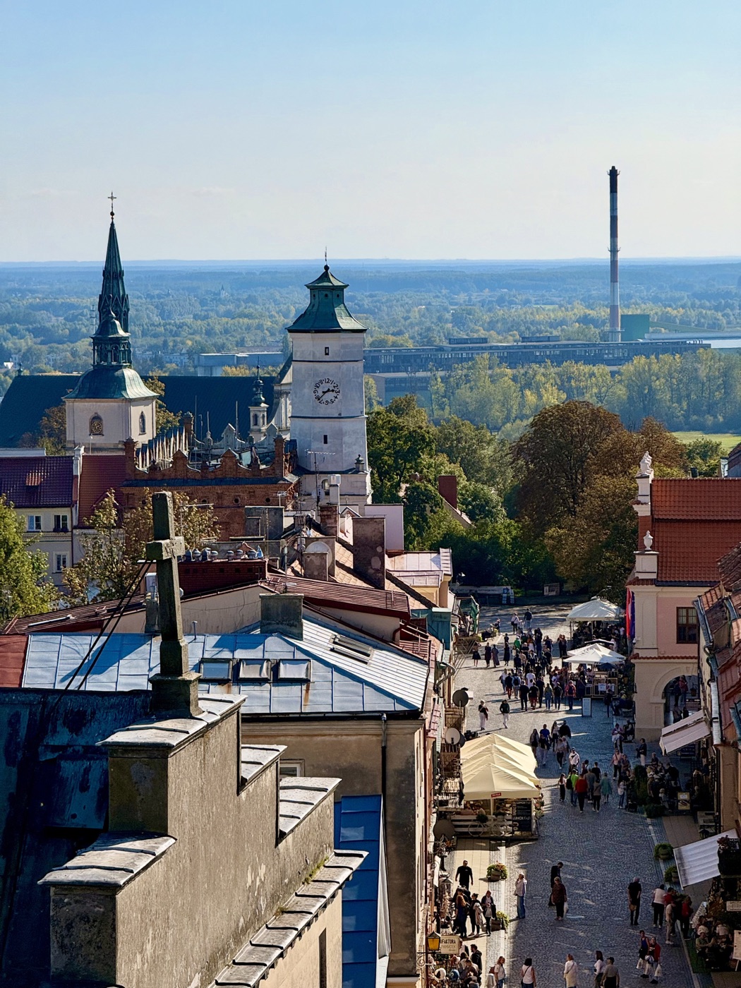 Widok z góry na brukowaną ulicę z ogródkami pod jasnymi parasolami, ceglany ratusz z attyką i białą wieżą z zegarem, z lewej wieża z iglicą, w tle zielona dolina i lasy po horyzont